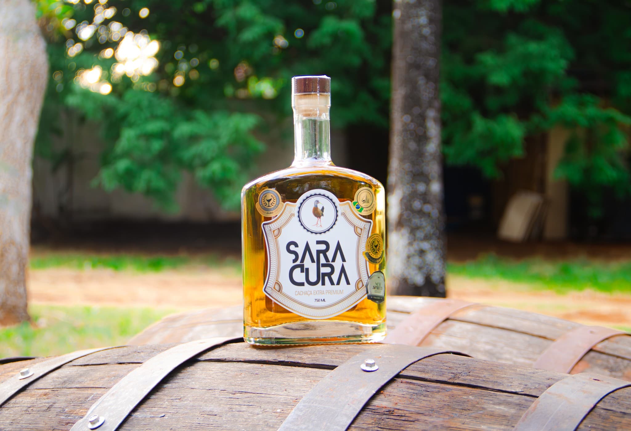 Garrafa de cachaça Saracura sobre um barril de madeira, com árvores ao fundo.