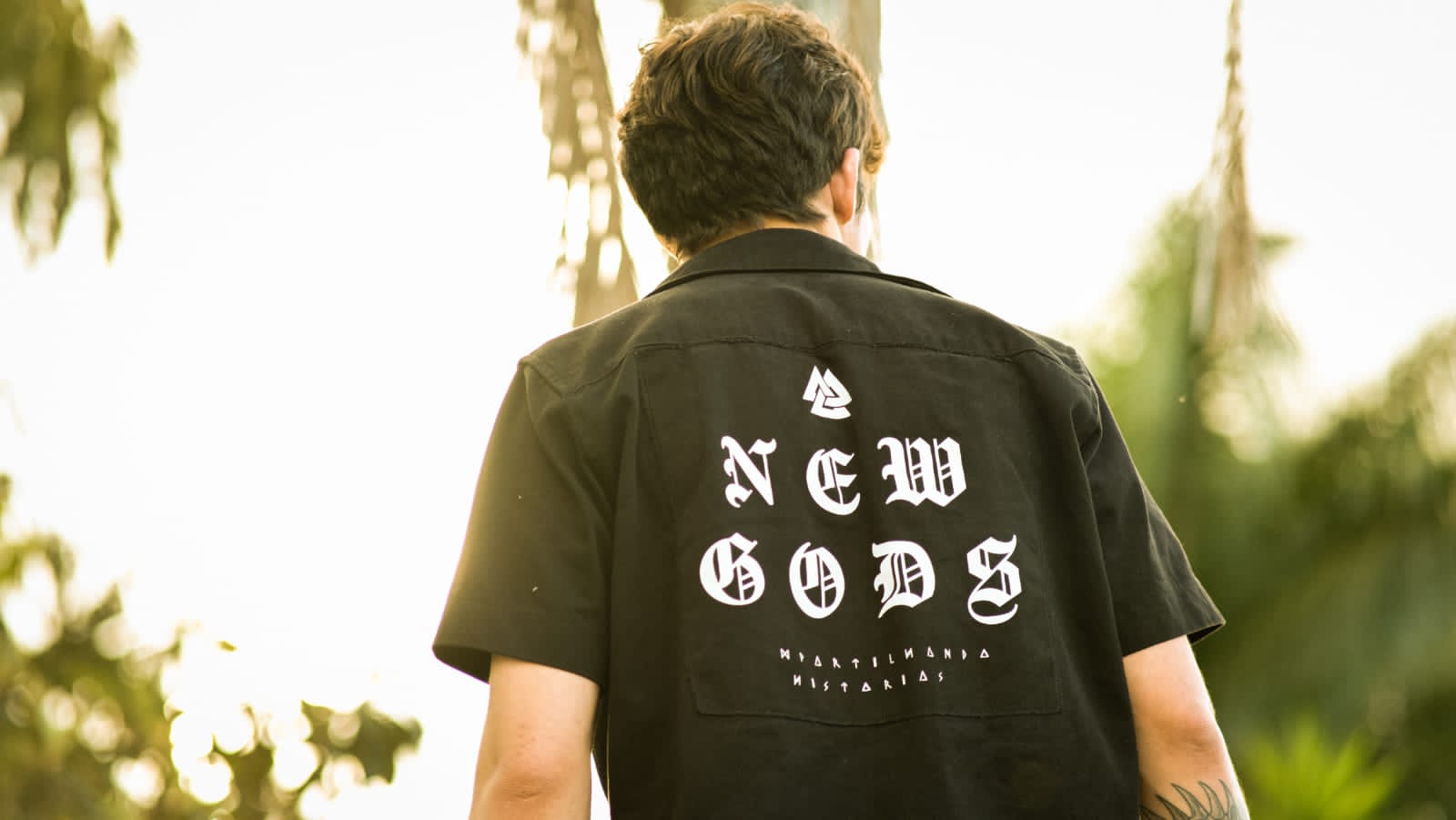 Homem de costas com camisa preta bordada com o nome NEW GODS, entre palmeiras ao sol.