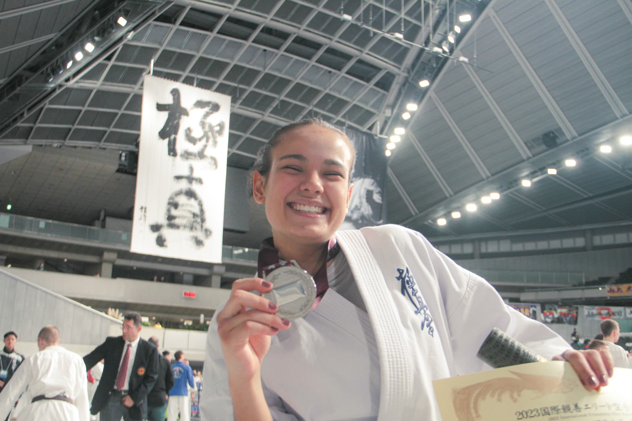 Atleta de quimono sorri e mostra uma medalha e um certificado no ginásio.