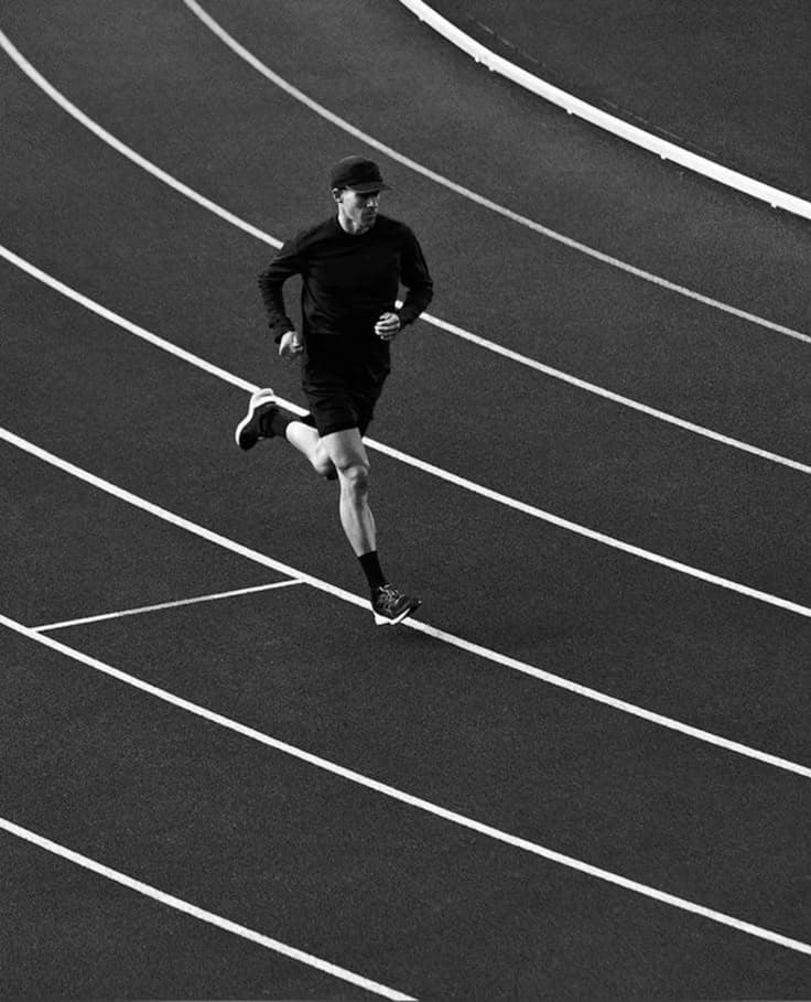 Corredor em uma pista de atletismo, fotografado de cima em preto e branco.