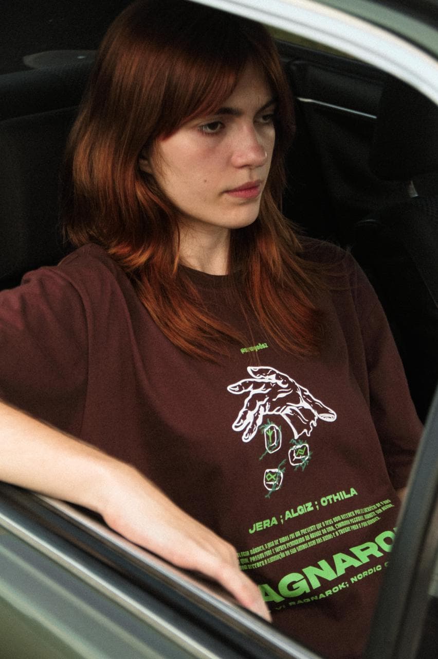 Jovem ruiva com camiseta marrom estampada NEW GODS, saindo de um carro.