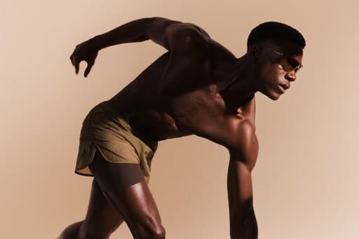 Atleta sem camisa e de óculos escuros em posição de largada, sobre fundo bege.