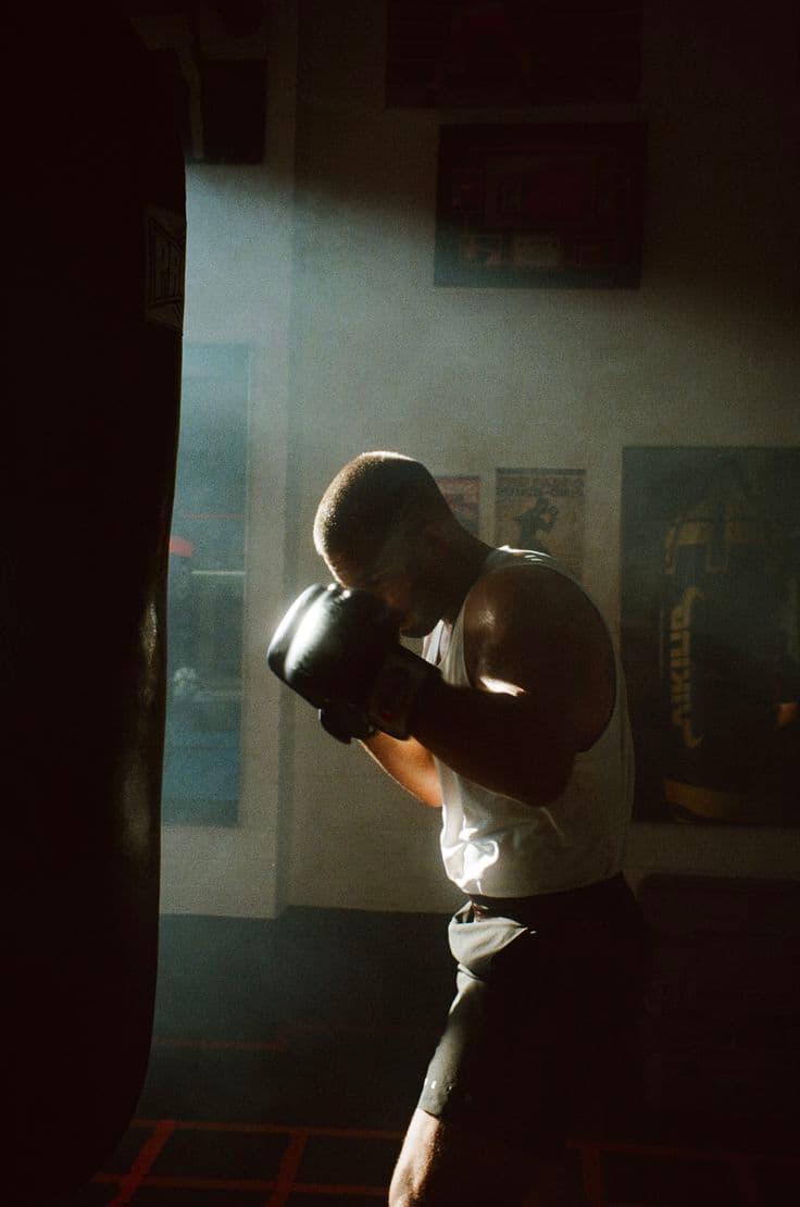 Boxeador de luvas diante de um saco de pancadas, iluminado pela luz lateral de uma janela.