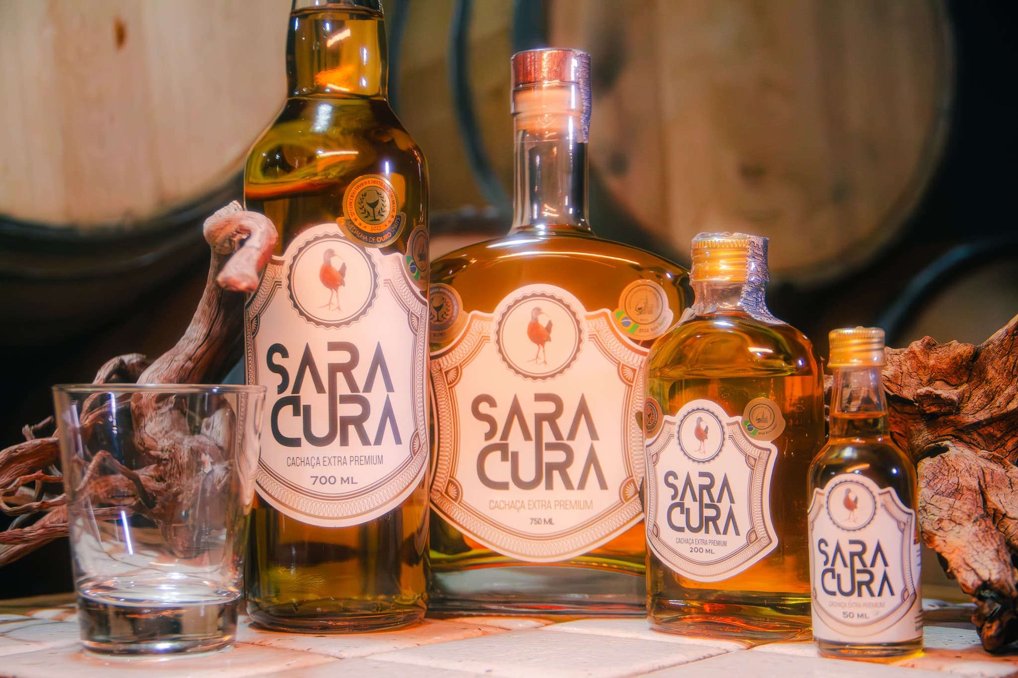 Garrafas de cachaça Saracura de vários tamanhos e um copo, diante de barris de madeira.