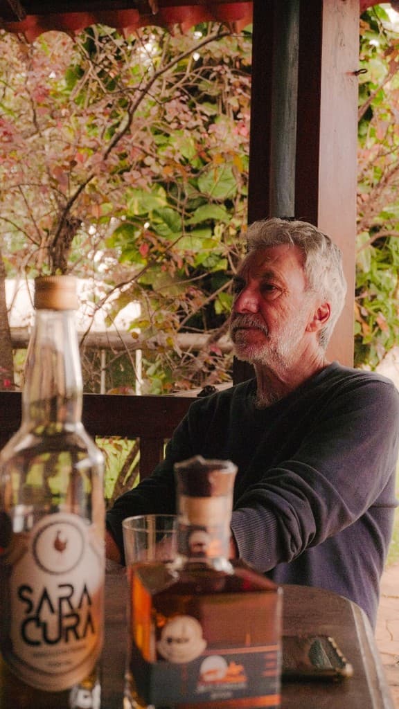 Homem grisalho sentado à mesa em uma varanda, ao lado de garrafas de cachaça Saracura.