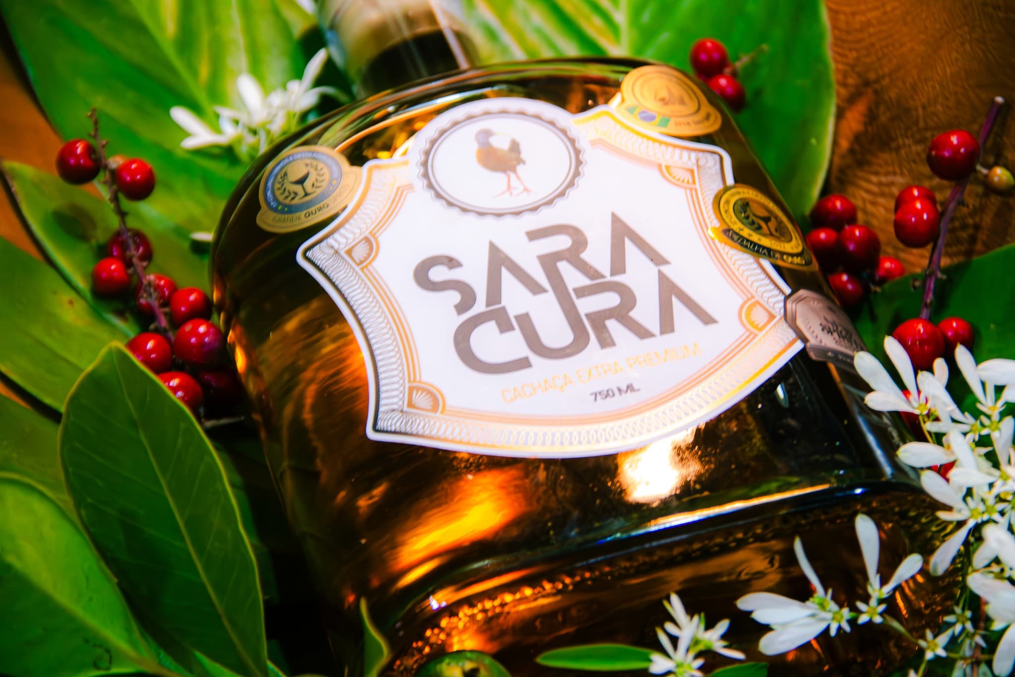 Garrafa de cachaça Saracura deitada entre folhas, frutos vermelhos e flores brancas.